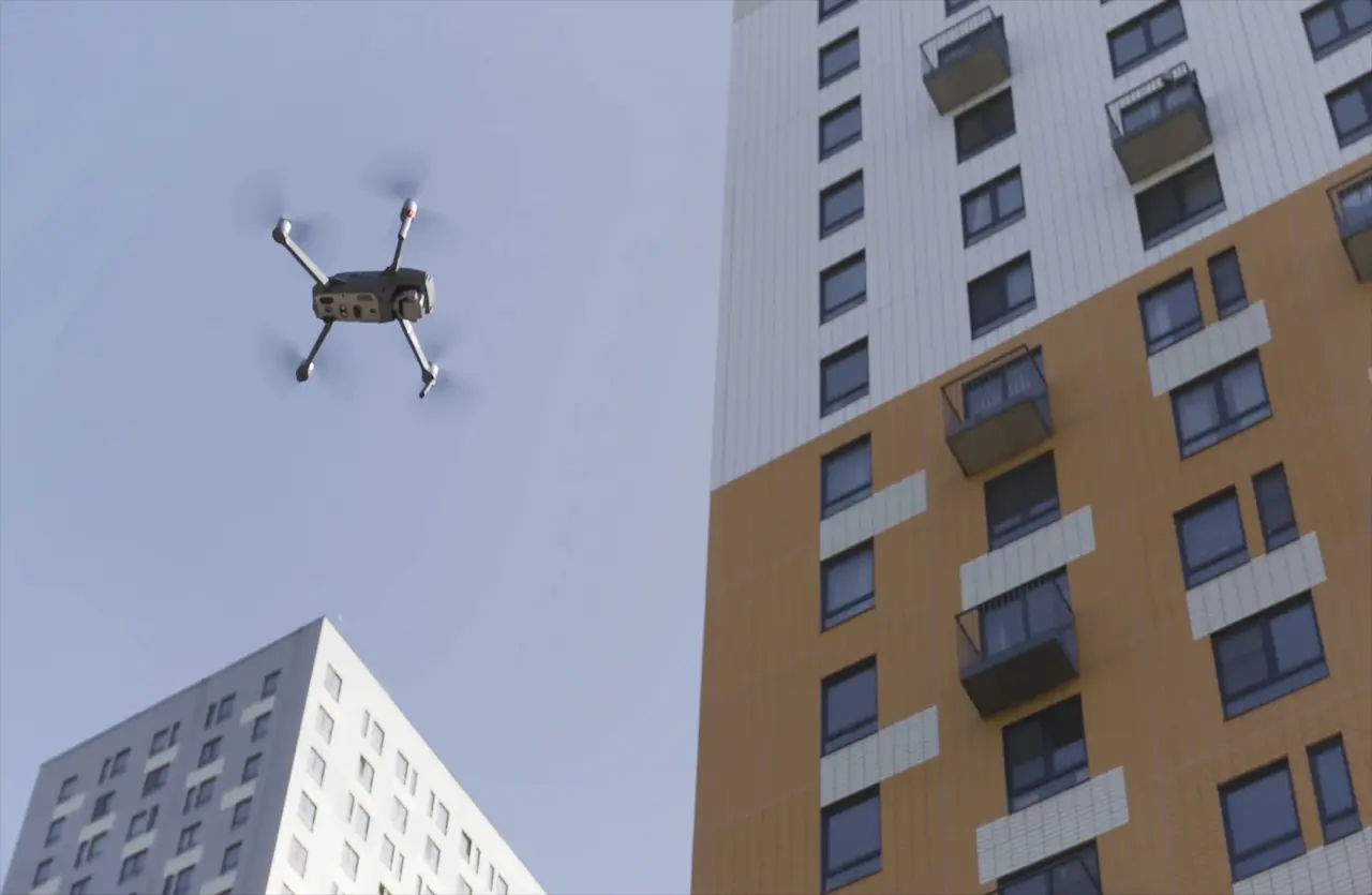 A drone hovers close to a modern high rise building, capturing views of its glass exterior and surrounding cityscape. The small aircraft flies near the structure, showing its sleek design, reflective windows, and the urban environment around it as it records footage.